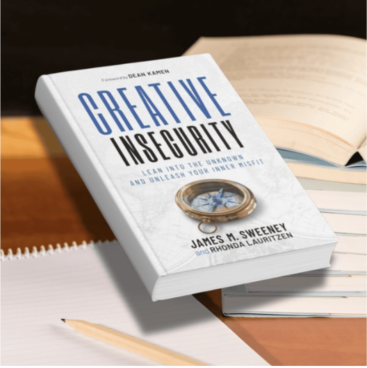 book A hardcover copy of the book Creative Insecurity, written for entrepreneurs by serial entrepreneur James M. Sweeney and Rhonda Lauritzen, floats on a desk beside open notebooks.